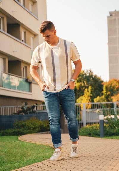Camisa de manga corta color crema con rayas verticales grises, combinada con jeans azules y zapatillas blancas, sobre un camino adoquinado en un entorno de jardín.