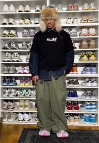 Homme portant un chapeau en fourrure, un t-shirt noir sur une chemise bleue, un pantalon vert ample et des baskets avec des accents roses, debout devant une grande étagère remplie de baskets.