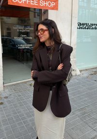 Femme aux longs cheveux bruns portant un blazer marron oversize et des lunettes orange, debout les bras croisés devant un café sur un trottoir en ville.