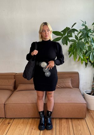 Vestido de punto negro con mangas largas tipo globos, cinturón plateado con detalles circulares, bolso negro acolchado y botas negras gruesas.