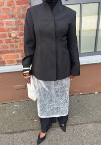 Czarny żakiet o dopasowanym kroju z wysokim kołnierzem, zestawiony z przezroczystą białą koronkową spódnicą, czarnymi spodniami i szpiczastymi czarnymi butami.