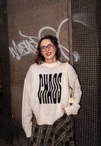 Pull crème surdimensionné avec le mot « CHAOS » en noir, associé à une jupe à carreaux et un sac de couleur claire. Sur un fond carrelé.