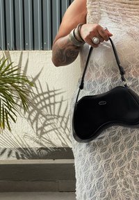 Schwarze Lederhandtasche mit einer einzigartigen, organischen Form, Reißverschluss und einem verstellbaren Trageriemen. Kleid mit Spitzenüberzug und Palmen-Schatten im Hintergrund.