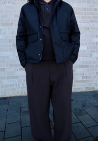 Must värvitud veekindel jope, millel on tõmblukk ja nööbid, koos tumesinise polo särgi ja laiade pruunide pükstega.