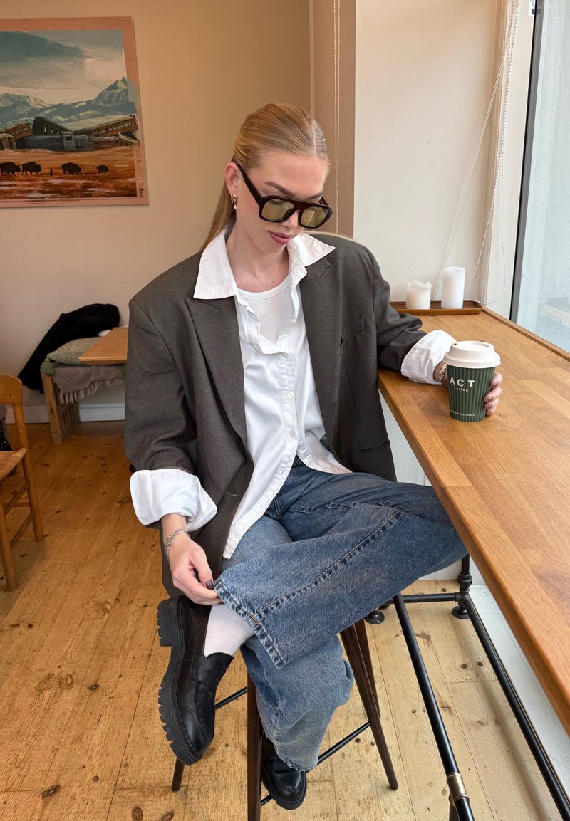 Blazer grigio oversize sopra una camicia bianca, jeans di denim blu e scarpe nere chunky. Tene in mano un bicchiere di carta verde per il caffè. Sfondo in legno e bianco.