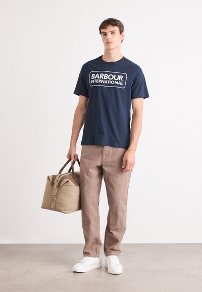 Camiseta de algodón azul marino con "BARBOUR INTERNATIONAL" impreso en blanco. Acompañada de pantalones marrón claro y una bolsa de lona color tostado. Zapatillas blancas.