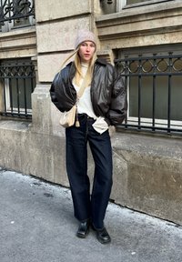 Καφέ μποφάν μπουφάν πάνω από ένα λευκό ριγωτό πουλόβερ, σκούρες μπλε φαρδιές τζιν παντελόνες, μαύρες χοντρές μπότες και ένα ανοιχτό ροζ πλεκτό σκουφάκι.