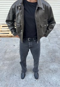 Veste en cuir verte avec des accessoires en argent, t-shirt noir, jeans gris foncé et chaussures noires. La veste dispose d'une poche avant et d'une fermeture éclair.
