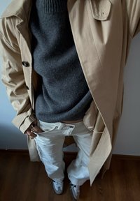 Άτομο που φοράει μπεζ καμπαρντίνα, σκούρο γκρι πουλόβερ, άσπρο παντελόνι και γκρι-άσπρα αθλητικά παπούτσια, στέκεται σε ξύλινο πάτωμα.