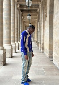 Pull en tricot avec un motif ondulé bleu et blanc, associé à un jean bleu clair déchiré. Baskets bleues et lunettes de soleil argentées notées.