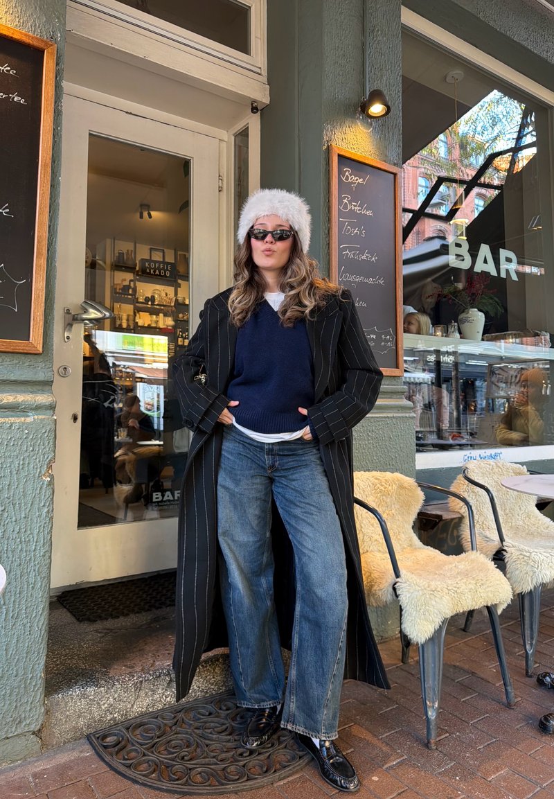 Manteau à fines rayures noires, pull marine, jean large, chapeau en fourrure et lunettes de soleil. Chaussures : mocassins noirs polis. Lieu : entrée d'un café.