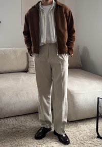 Homme debout à l'intérieur portant une veste marron, une chemise rayée sur un t-shirt blanc, un pantalon beige et des chaussures noires sur un sol recouvert de moquette.