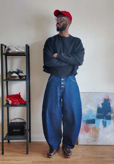 Hombre con gorra roja, sudadera negra recortada, vaqueros azules holgados y zapatos marrones, de pie con los brazos cruzados junto a un zapatero y una pintura abstracta.
