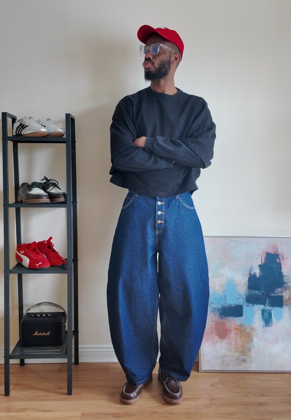 Man wearing red cap, black cropped sweatshirt, wide blue jeans, and brown shoes stands with arms crossed next to shoe rack and abstract painting.