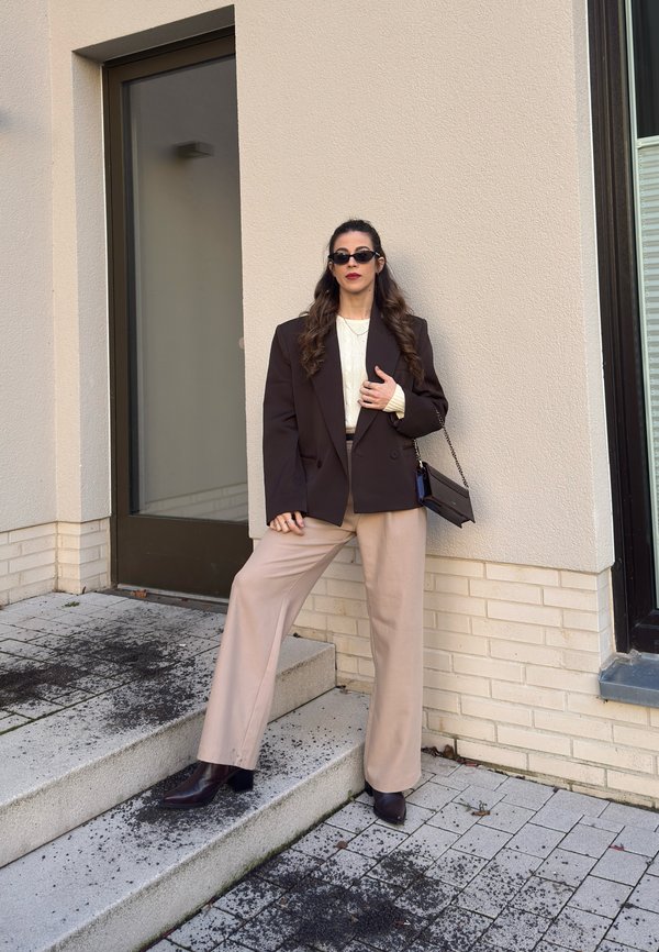 Vrouw met een donkere oversized blazer, crèmekleurige trui, beige wijde broek, donkere laarzen, zonnebril en een zwarte handtas in haar hand, staand op buitentrap.