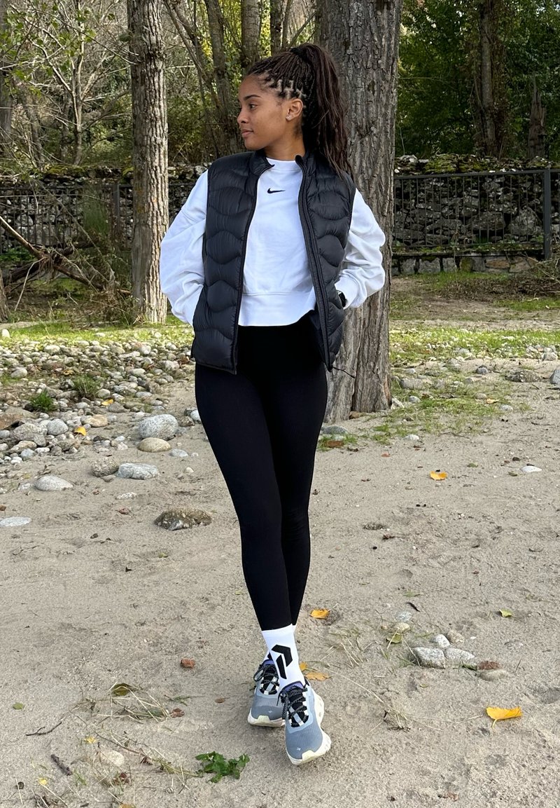 Μαύρο γιλέκο πούφερ πάνω από λευκό πουκάμισο με μακριά μανίκια, μαύρες κολάν, και γκρι αθλητικά παπούτσια με λευκές καλτσες με σχέδια, σε εξωτερικό χώρο.