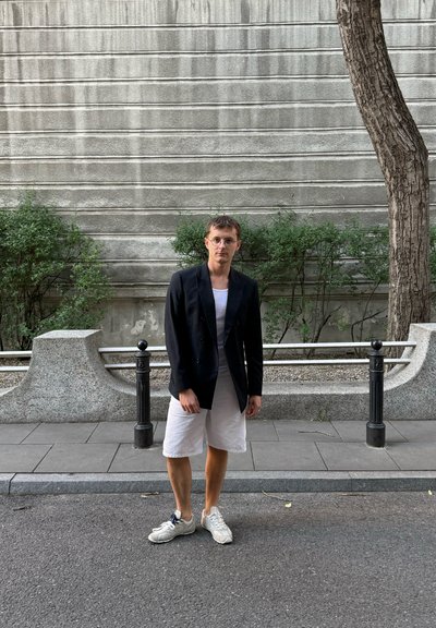 Blazer negro de doble botonadura, camiseta gris, pantalones cortos blancos y zapatillas claras. Pared texturizada gris y vegetación en el fondo.