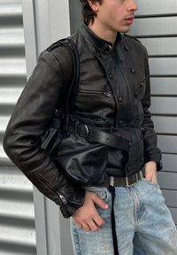 Ung mand iført sort læderjakke og lyseblå jeans, bærende på en sort læderhåndtaske, stående foran en baggrund af metalpaneler.