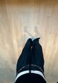 Svarta byxor med slim fit och texturerad tyg, prydda med ett dekorativt bälte av flerfärgade pärlor, placerade på en ljus träplatta.