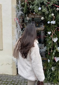 Femme aux longs cheveux bruns portant une veste polaire blanche, regardant une porte festive décorée de branches de pin, de cannes en sucre d'orge et d'ornements blancs.