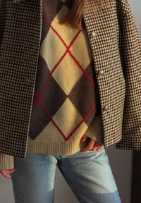 Personne portant une veste marron à carreaux sur un pull beige à losanges avec des diamants rouges et marron foncé, assortis à un jean bleu clair.