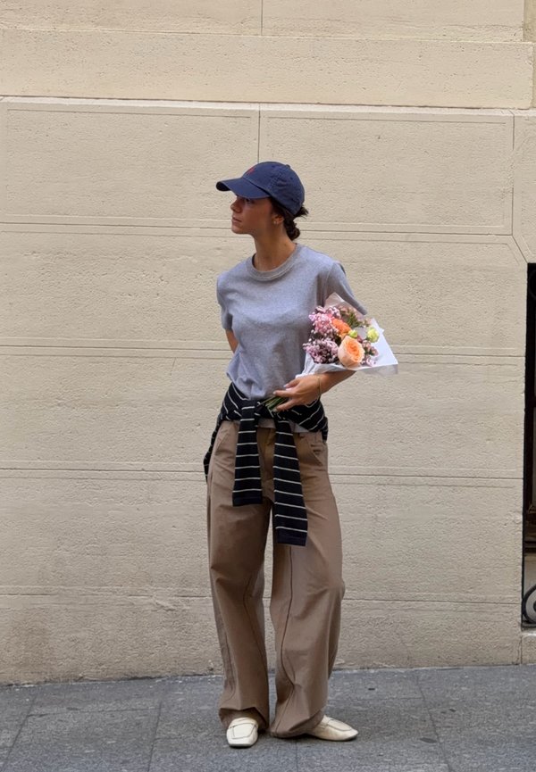 Femme portant une casquette, un t-shirt gris et un pantalon large, se tient debout avec un bouquet de fleurs devant un mur beige.
