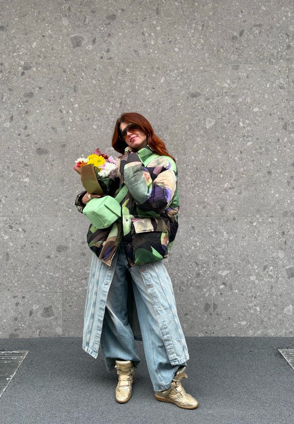 Kamuflažna puhovka v zelenih in vijoličnih odtenkih, preveliki svetlo modri jeans hlače, zlate superge, drži šopek cvetja, sončna očala.