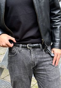 Czarny pleciony skórzany pasek z srebrną klamrą noszony na ciemnoszarych dżinsach i czarnej koszuli, uzupełniony czarną skórzaną kurtką.