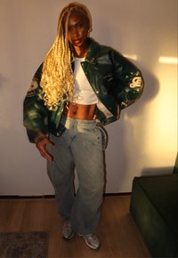 Zelená kožená bomber bunda s emblémy, bílý crop top, světle modré volné džíny a šedé tenisky. Dlouhé blond pletené vlasy.