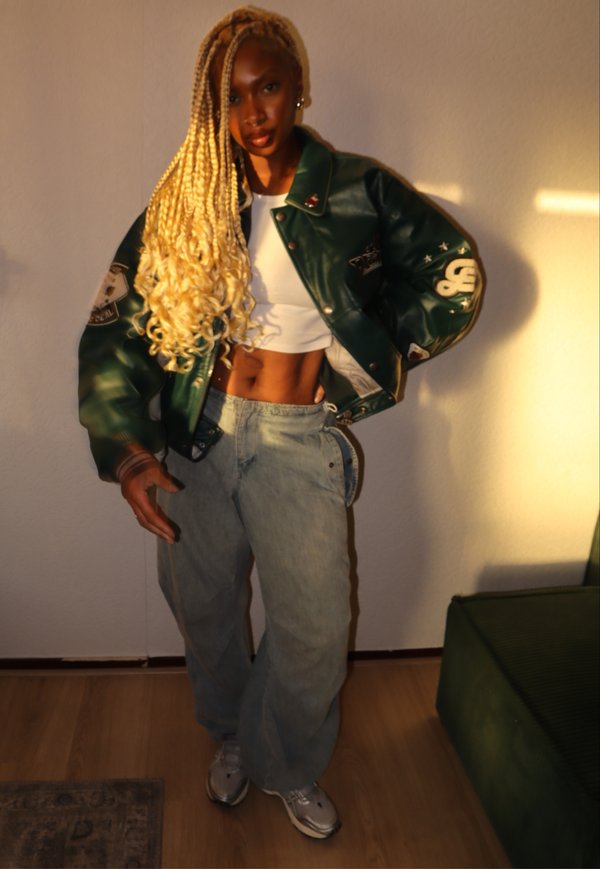 Groene leren bomberjack met emblemen, witte crop top, lichtblauwe baggy jeans en grijze sneakers. Lange blonde gevlochten haren.