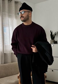 Pull côtelé bordeaux, pantalon large noir, et un manteau noir porté sur le bras. Chaise en bois et plante d'intérieur en arrière-plan.