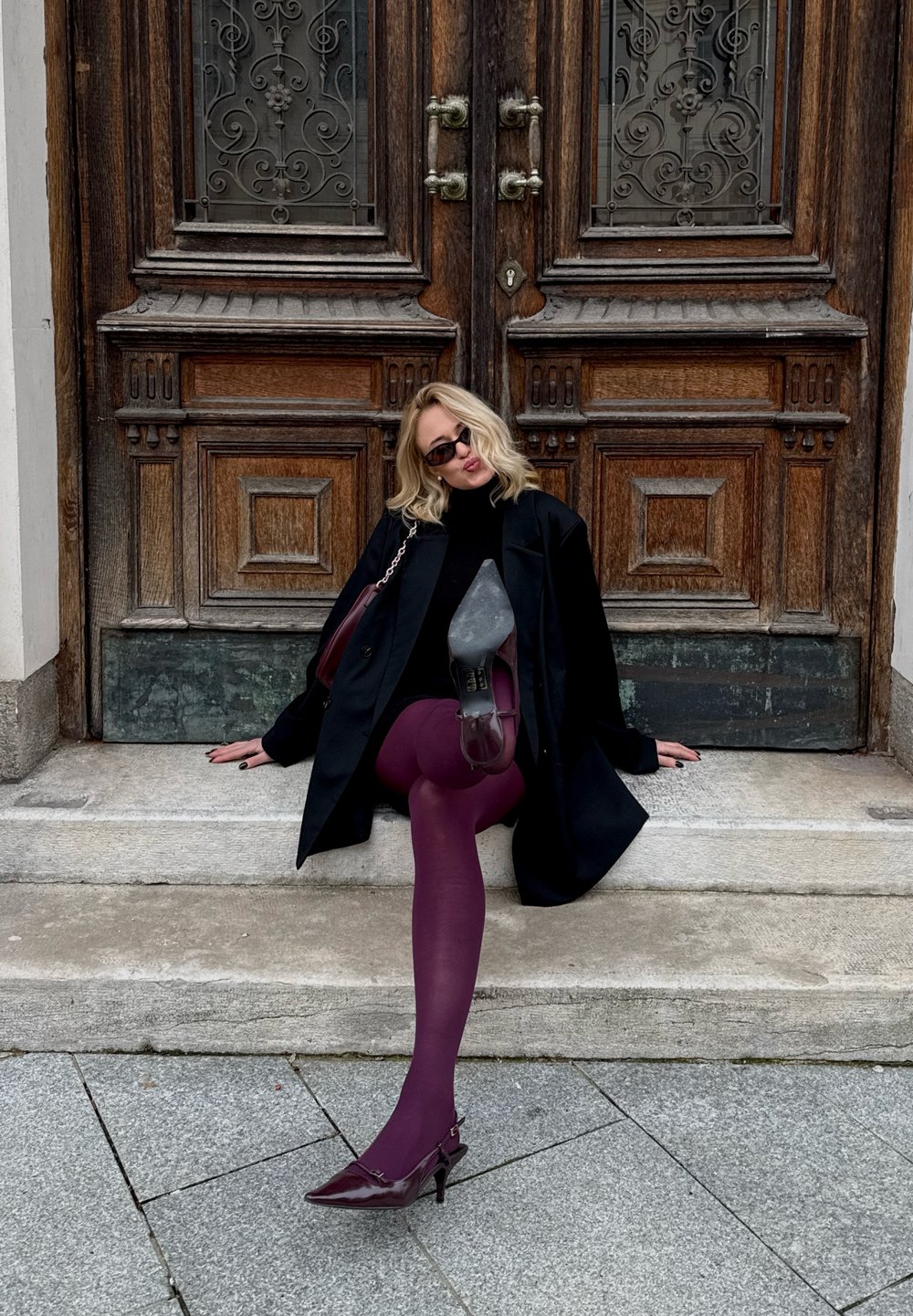 Black coat, black top, and purple tights; pointed burgundy heels; sitting on stone steps with ornate wooden doors behind.
