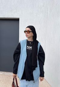 Τζιν γιλέκο πάνω από μαύρη φούτερ με κείμενο "NIKE", συνδυασμένο με μαύρη μαντίλα και μεγάλα πορτοκαλί γυαλιά ηλίου. Κρατάει μια μπορντό τσάντα.