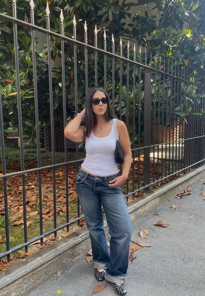 Top blanco sin mangas, jeans sueltos azul oscuro y zapatos negros con lunares. Bolso negro. Valla de metal y vegetación al fondo.