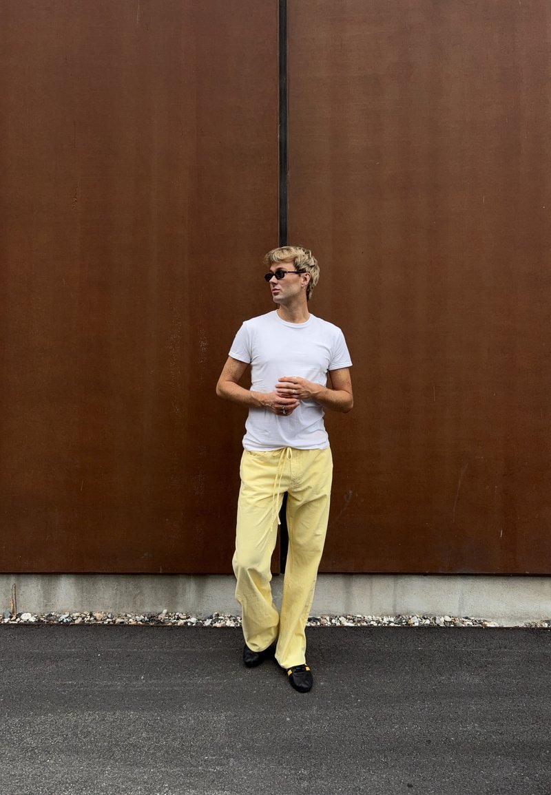 Homme portant des lunettes de soleil, un t-shirt blanc et un pantalon jaune, debout contre un grand mur métallique marron rouillé sur un sol en asphalte.