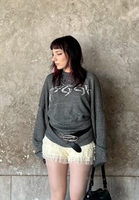 Grijze oversized trui met witte grafische print, gelaagd over lichtgele frivole shorts, gecombineerd met een zwarte riem en handtas, tegen een textuur achtergrond.