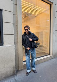 Jauns pieaugušais, valkājot melnu rāvējslēdzēja jaku, tumšu šalli ar bārkstīm, gaiši zilas platas džinsas un tumšas kurpes, stāv pie veikala loga.