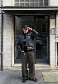 Zwarte leren bomberjack, grafisch zwart T-shirt, bruine wijde ribfluwelen broek, zwarte pet, sleutelhanger zichtbaar, staande buiten een gebouw.