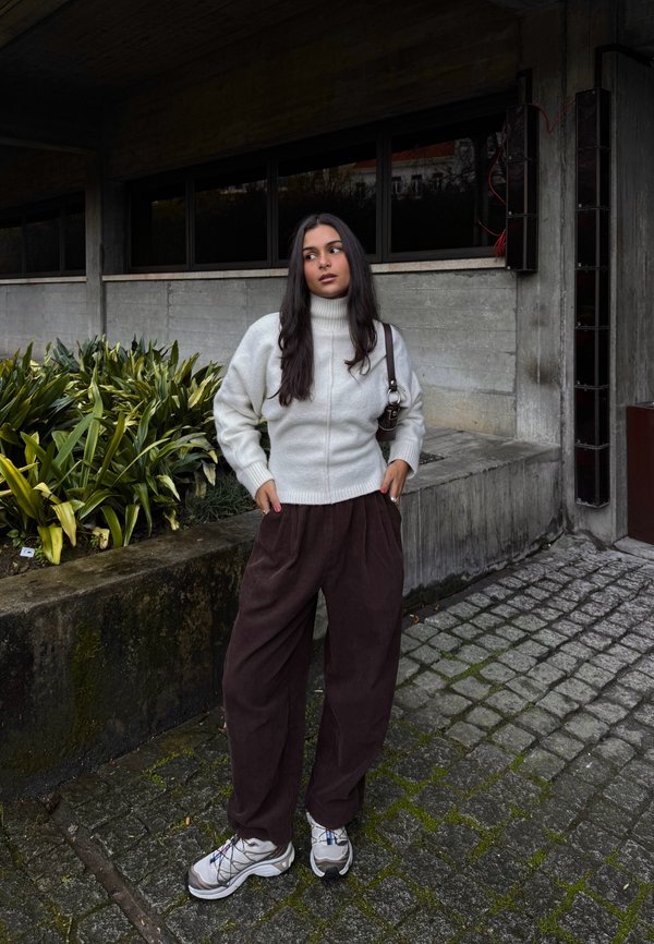 Femme portant un pull à col roulé blanc et un pantalon marron, debout les mains dans les poches près de plantes vertes et de murs en béton en plein air.