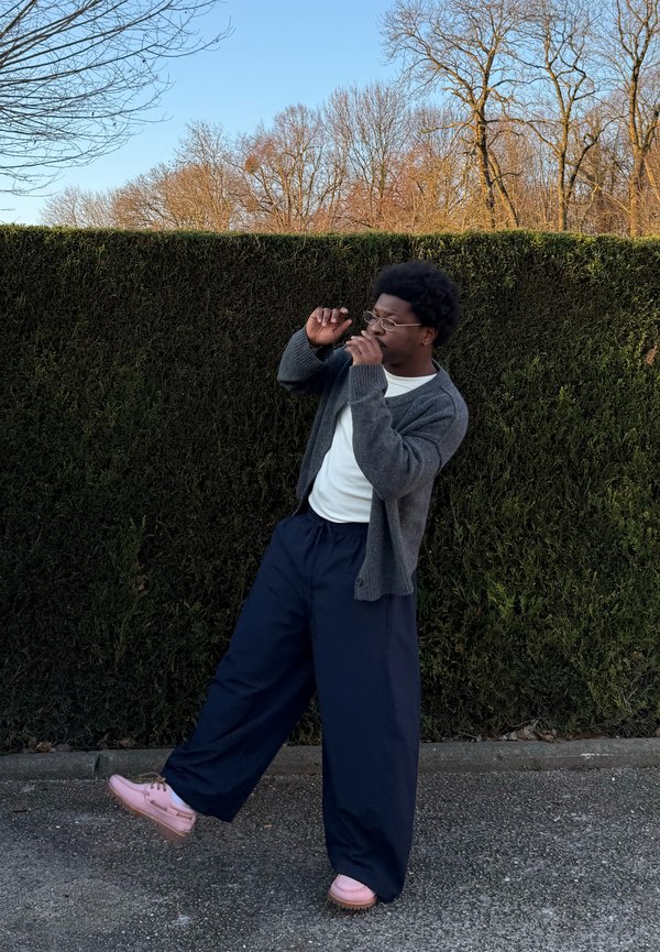 Young person wearing glasses, gray cardigan, white shirt, navy wide pants, and pink shoes poses with one leg raised in front of a tall green hedge.