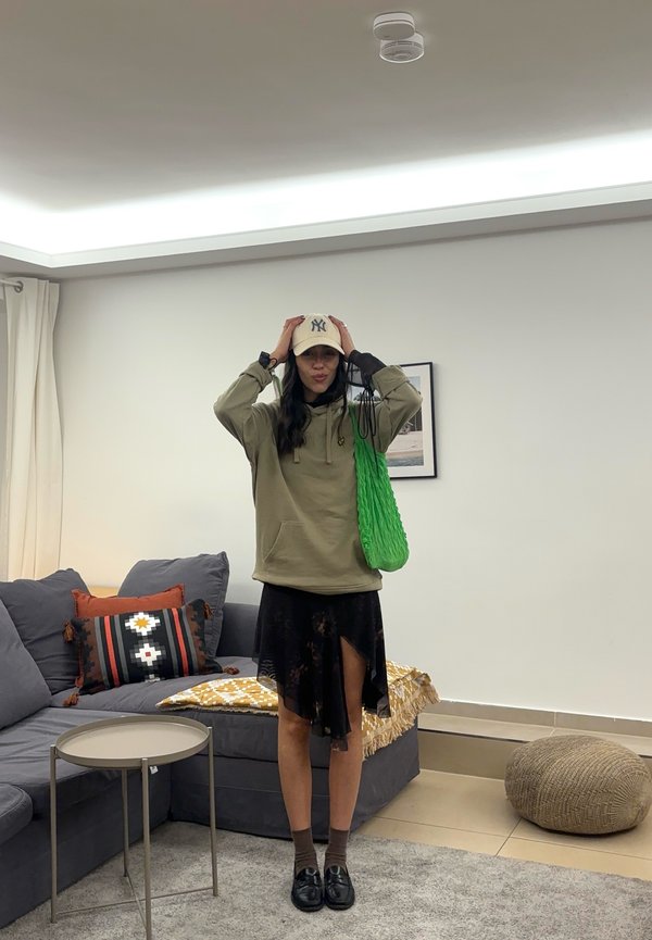 Oliven grønt hoodie, sort nederdel med en side-slids, beige kasket, grøn flettet taske, mørke sokker og sorte sko. Indendørs stemning med grå sofa.
