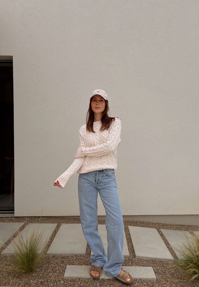Suéter de punto trenzado crema, jeans de pierna ancha azul claro y sandalias beige. El sombrero presenta un logo sutil. Texturas suaves y corte relajado.