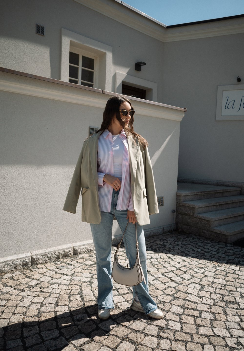 Veste oversize beige clair, chemise rayée rose et blanche, jean évasé bleu clair et sac à épaule gris clair. Sol en pavés.