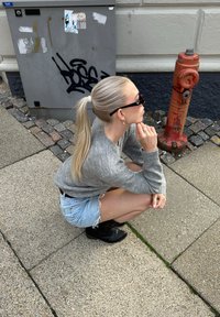 Siv svitek z pletenine, svetlo modri raztrgani jeans kratki hlače, črne gleženjske škornje in sončna očala; postavljena v čepenju blizu rdeče hidrantne naprave in grafitov.