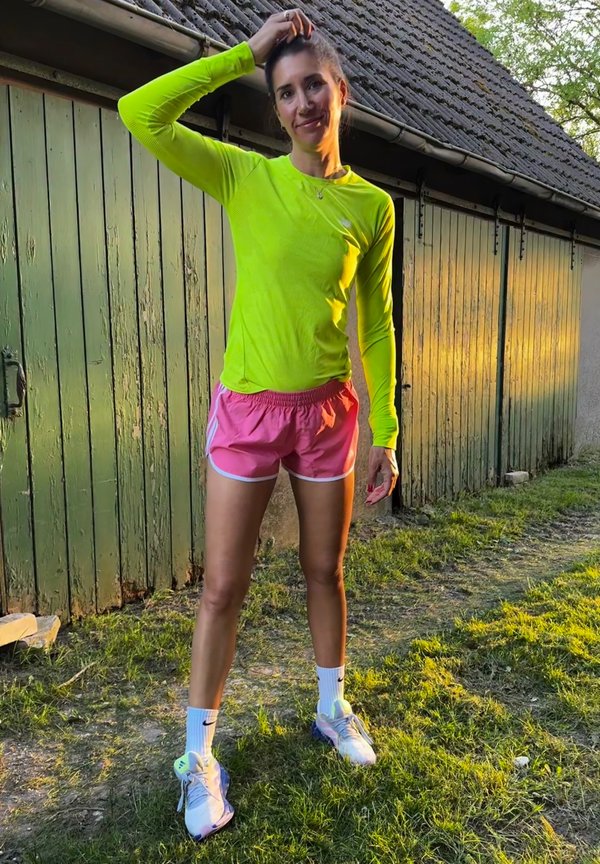 Neon zelené tričko s dlhým rukávom, ružové kraťasy s bielym lemovaním, biele ponožky s logom a svetlo modré športové topánky, stojace na tráve.