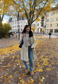 Nuori nainen beigenvärisessä takissa ja farkuissa seisoo syksyn lehtien peittämällä kaupunkialueen jalkakäytävällä, hymyilee ja katsoo sivulle.