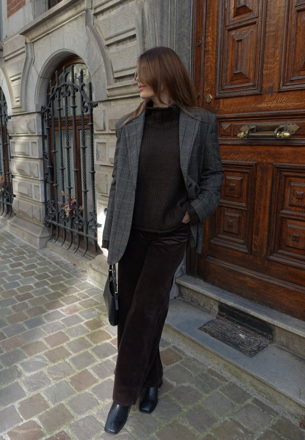 Grå rutete blazer, brun strikket genser, brune kordfløyelsbukser, svarte ankelstøvler og en svart veske foran en tredør og brosteinsbelagt fortau.