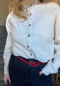 Balts adīts kardigans ar koka pogām, kombinēts ar tumši zilām džinsa biksēm un sarkanu jostu. Apģērbs demonstrē gludu faktūru un vienkāršu dizainu.