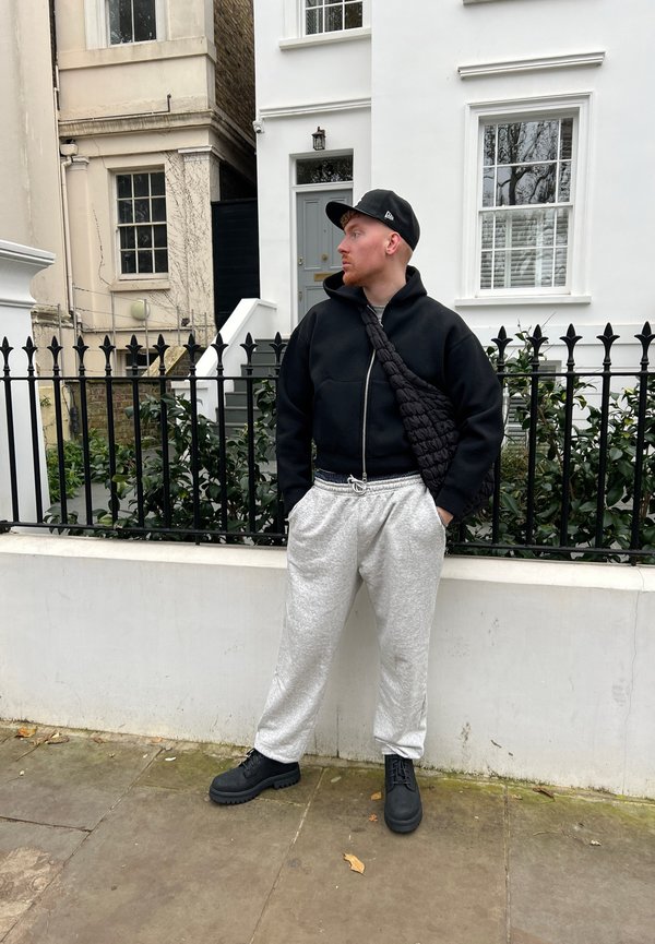 Tenue monochrome avec un sweat à capuche noir zippé, un jogging gris et des bottes noires à lacets. Un sac en bandoulière texturé est porté en diagonale.