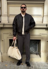 Mann trägt dunkle Sonnenbrille, schwarze Lederjacke, graue Hose, schwarze Schuhe, hält eine große beige Tragetasche, steht an einer beigen Wand.